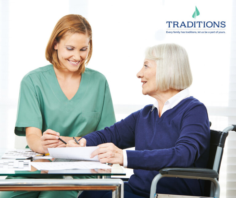 A senior woman sitting in a wheelchair signing documents as her female caregiver sits beside her with the Traditions Management logo in the upper righthand corner of the image