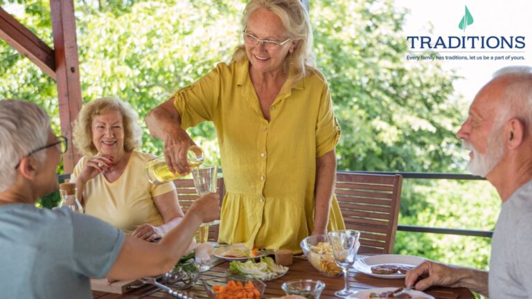 A senior woman pouring juice into another senior woman's cup outdoors at a picnic table with two other seniors sitting at the table with the Traditions Management logo in the top righthand corner of the image