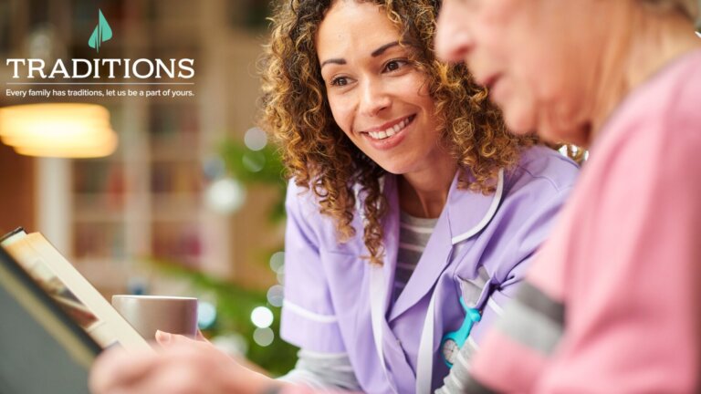 Close up of a caregiver wearing purple scrubs smiling as she looks at a memory book her female patient is looking down at with the Traditions Management logo in the top lefthand corner