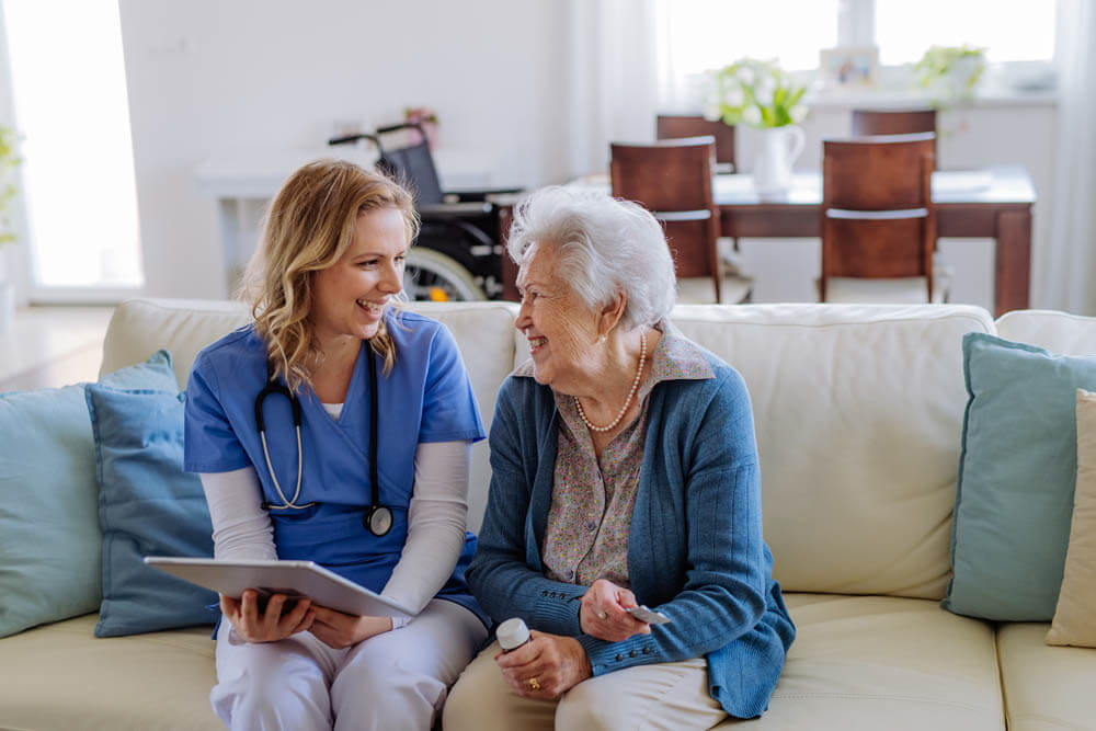 Female nurse sitting on a sofa with a memory care patient, laughing and conversing, at Traditions of Hilliard in Hilliard, OH.